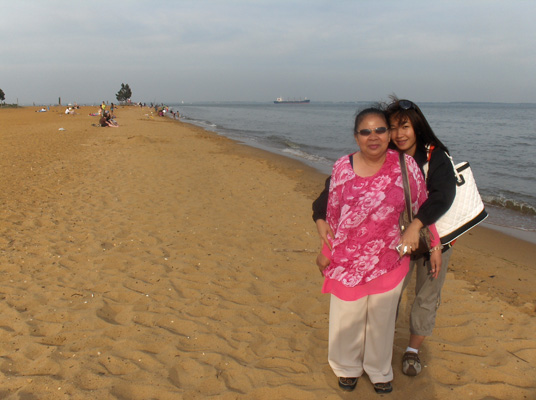 Ann hugs Nanta from behind her.  They are standing on the beach.  Barely visible on the other side of the water is a long bridge.